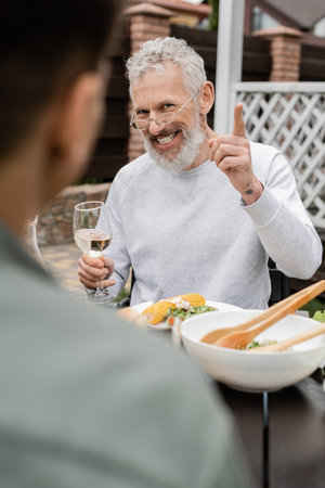 cheerful middle aged man looking son and gesturing during bbq party, blurred foreground, sitting on backyard of summer house, spending time together, eating grilled bbq food, parents day conceptの写真素材