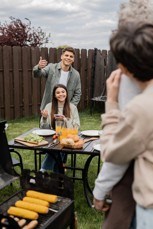 parents day celebration, happy siblings looking at father and mother on blurred background, inviting to table, gesturing, family bbq, grill party, preparing food on grill bbq, modern parentingの写真素材