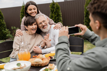 Smiling teenage girl hugging middle aged parents near blurred brother taking photo on smartphone near bbq food during parents day celebration at backyard, special day for parents conceptの写真素材