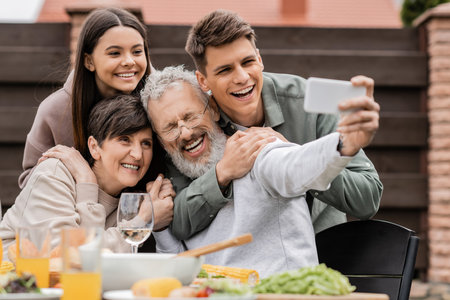 Cheerful kids hugging middle aged parents while taking selfie on smartphone together during barbeque party and parents day celebration at backyard in june, happy parents day conceptの写真素材