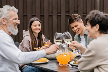 Smiling siblings sitting near tasty bbq food and blurred middle aged parents toasting with wine during parents day celebration at backyard, family love and unity concept, tradition and celebrationの写真素材
