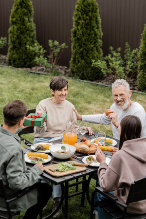 Young son giving gift box to cheerful middle aged mother near sister and father while celebrating parents day and family bbq party at backyard in june, cherishing family bonds conceptの写真素材