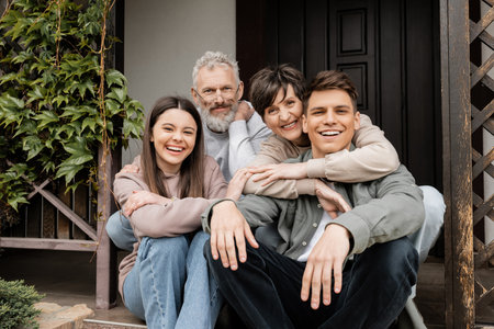 Positive middle aged parents hugging children and looking at camera while sitting on porch of house...