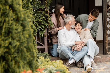 Smiling middle aged parents holding hands while sitting on porch of house near kids and celebrating...