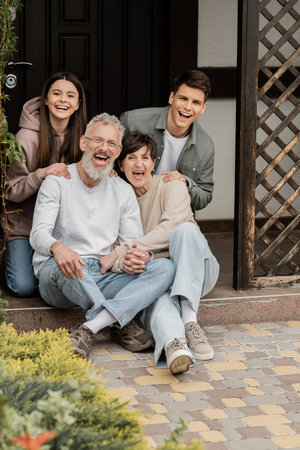 Excited and cheerful middle aged parents holding hands and looking at camera near children on porch...