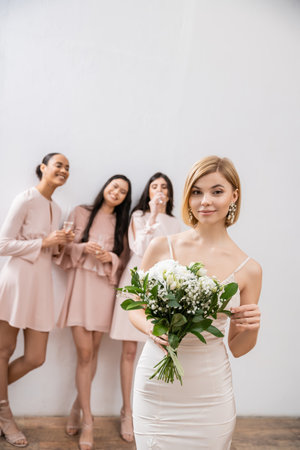 beautiful bride in wedding dress holding bridal bouquet, standing near blurred interracial bridesmaids on grey background, happiness, special occasion, blonde and brunette womenの写真素材
