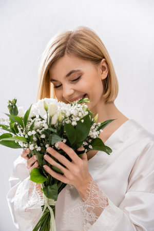 white flowers, happiness, cheerful bride with blonde hair standing in white silk robe and smelling bridal bouquet, young woman, beautiful, excitement, feminine, blissful, portrait, grey backgroundの写真素材