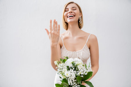 special occasion, gorgeous blonde bride in wedding dress holding bouquet and showing engagement ring, white flowers, bridal accessories, happiness, grey backgroundの写真素材