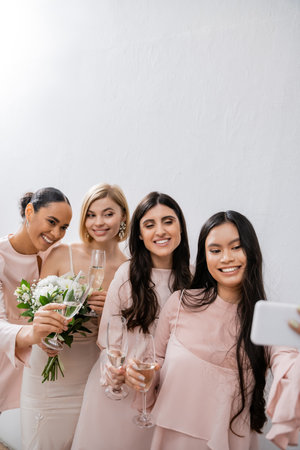 four women, cheerful blonde bride and her interracial bridesmaids taking selfie together, happiness, champagne glasses, bridal bouquet, wedding dress, bridesmaid gown, brunette and blonde womenの写真素材