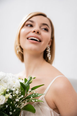 special occasion, beautiful, blonde bride in wedding dress holding bouquet on grey background, white flowers, bridal accessories, happiness, feminine, blissful, looking away, joyの写真素材