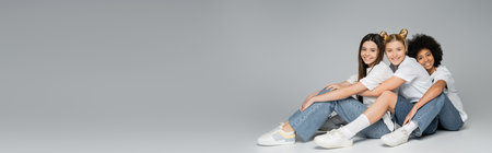Full length of positive teen multiethnic girlfriends in casual t-shirts and jeans looking at camera while sitting on grey background, multiethnic teen models concept, banner with copy spaceの写真素材