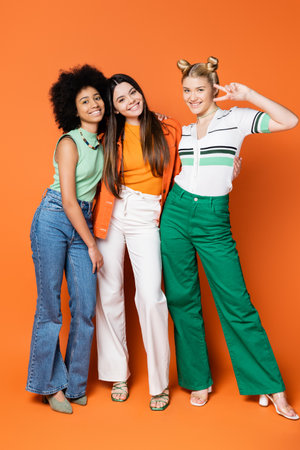 Full length of positive blonde teenager showing victory gesture while posing with stylish multiethnic girlfriends and standing on orange background, cool and confident multicultural teenage girlsの写真素材