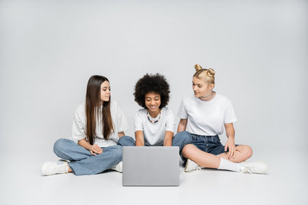Smiling teen african american girl using laptop near girlfriends in white t-shirts and jeans and sitting together on grey background, teenagers bonding over common interestの写真素材