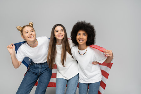 Teenage and multiethnic girlfriends in white t-shirts and jeans smiling away while holding american flag and standing on grey background, energetic teenage friends spending time, friendshipの写真素材