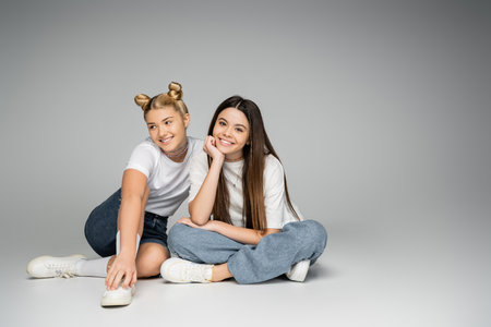 Cheerful and teen girlfriends in white t-shirts, sneakers and denim shorts smiling at camera while sitting together and posing on grey background, lively teenage girls concept, friendship and bondingの写真素材