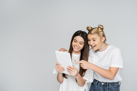 Smiling blonde teen girl in jeans and white t-shirt pointing with finger at digital tablet and hugging brunette friend isolated on grey, teenagers bonding over common interest, Touch Screenの写真素材
