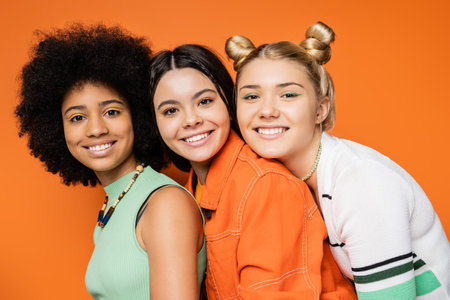Portrait of cheerful and multiethnic stylish teenage girlfriends with bold makeup wearing casual clothes and looking at camera together on orange background, stylish and confident posesの写真素材