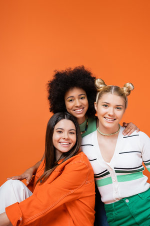 Smiling african american teenage girl with bright makeup hugging stylish girlfriends in casual outfits and posing together isolated on orange, cool and confident teenage girlsの写真素材