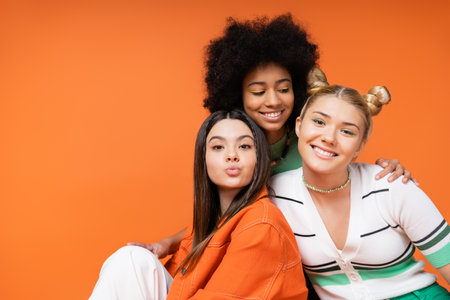 Cheerful african american teen girl with bright makeup hugging stylish blonde and brunette girlfriends while posing together isolated on orange, cool and confident teenage girlsの写真素材
