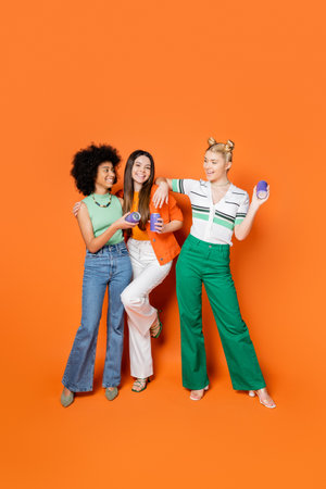 Full length of positive and multiethnic teenage girlfriends in stylish outfits holding canner drink and posing on orange background, trendy outfits and fashion-forward looksの写真素材