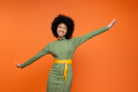 Cheerful and teen african american girl in stylish green dress and bold makeup winking and looking at camera while standing isolated on orange, teenage fashion and generation z conceptの写真素材
