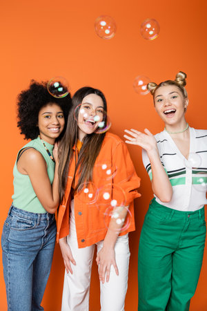 Soap bubbles near positive and interracial teenage girlfriends in casual outfits looking at camera...