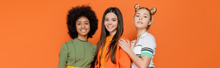 Trendy blonde teenage girl hugging multiethnic friends in stylish outfits while standing and posing together isolated on orange, trendy generation z concept, banner, friendship and companionshipの写真素材