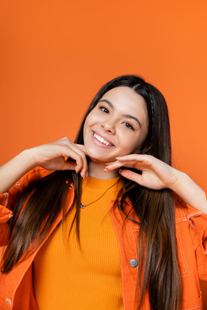 Portrait of positive and stylish brunette teenage model in denim jacket touching chin and smiling at camera while standing isolated on orange, cool and confident teenage girlの写真素材