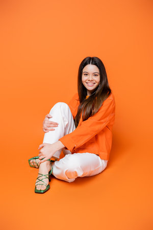 Full length of positive brunette teenager in heels and denim jacket in stylish outfit touching leg while sitting and posing on orange background, cool and confident teenage girl, gen z fashionの写真素材