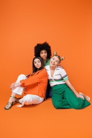 Full length of cheerful and multiethnic teen girlfriends in trendy casual outfits posing together and looking at camera on orange background, cool and confident multicultural teenage girlsの写真素材
