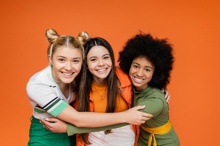 Portrait of positive and interracial teenage girlfriends in trendy outfits hugging each other and looking at camera while standing isolated on orange, trendy generation z conceptの写真素材