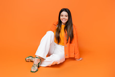 Fashionable and smiling brunette teenage girl in heels and denim jacket looking at camera while sitting and posing on orange background, cool and confident teenage girl, gen z fashionの写真素材