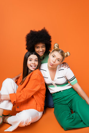 Positive and multiethnic teenage girlfriends in stylish casual outfits sticking out tongues and winking at camera while posing on orange background, cool and confident teenage girls, diverse racesの写真素材