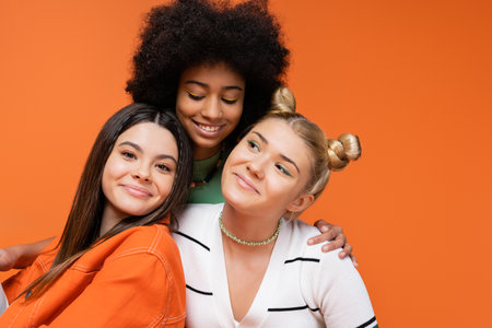 Smiling african american teen model with bright makeup hugging fashionable girlfriends and posing together isolated on orange, cool and confident teenage girls, diverse racesの写真素材
