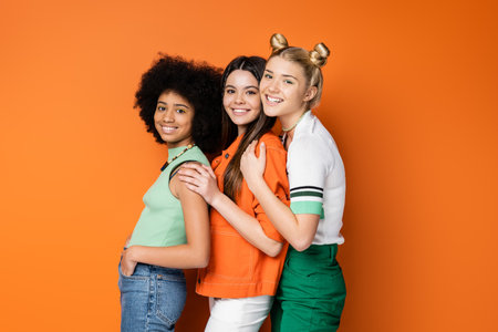 Trendy and smiling multiethnic teenage girlfriends with bold makeup wearing casual outfits while posing and looking at camera on orange background, stylish and confident posesの写真素材