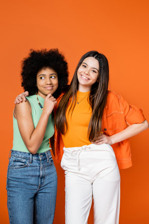 Smiling and stylish brunette teenager with colorful makeup hugging african american girlfriend in casual outfit isolated on orange, teen fashionistas with impeccable style concept, diverse racesの写真素材