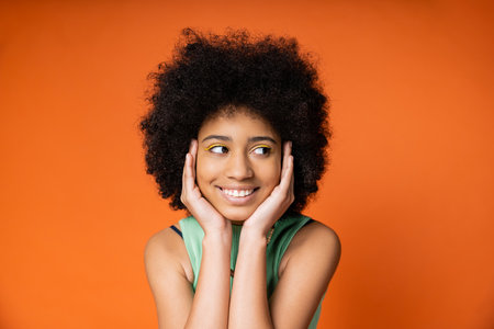 Pleased and stylish african american teenager with colorful makeup touching face and looking away while standing isolated on orange, trendy teenage girl expressing individualityの写真素材