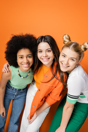 Portrait of multiethnic smiling teenage girls with colorful makeup and trendy outfits hugging while posing and looking at camera together on orange background, trendy and stylish hairstylesの写真素材