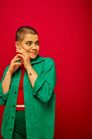 fashion and trends, tattooed, cheerful and short haired woman in green outfit posing on red background, looking away, generation z, youth culture, vibrant backdrop, individualityの写真素材