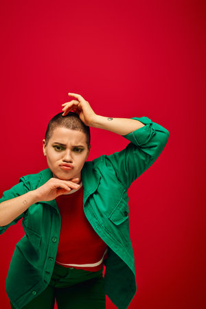 fashion and style, emotional and tattooed, short haired woman in green outfit pouting lips on red background, looking at camera, generation z, youth culture, vibrant backdrop, individualityの写真素材