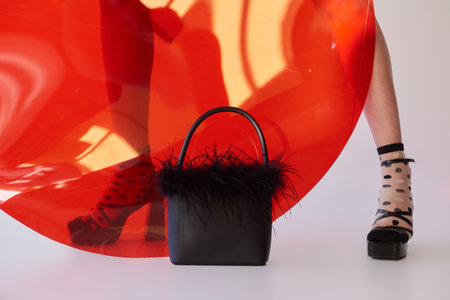 cropped view of woman in dotted socks and high heels standing near round shaped glass on grey background, feathered handbag, female legs, fashion statement, personal style, youthの写真素材