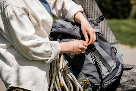 Cropped view of young tattooed female hiker in casual clothes checking backpack with equipment and...