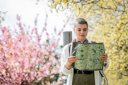 Young short haired female tourist in casual clothes with backpack looking at map while standing with blurred landscape at background, independent traveler embarking on solo journeyの写真素材