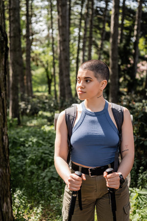 Young and confident short haired and tattooed female hiker with backpack holding trekking poles and looking away while standing in summer forest, hiking for health and wellness conceptの写真素材