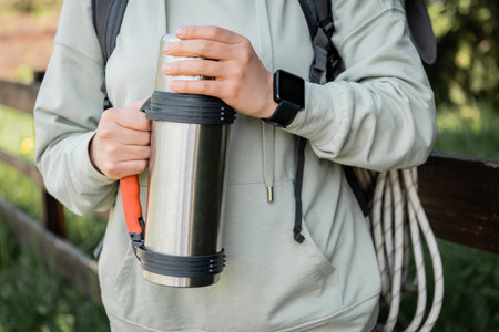 Cropped view of young woman tourist with backpack and fitness tracker holding flask while hiking with blurred nature at background, trekking through rugged terrain, summerの写真素材