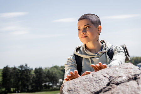 Portrait of smiling short haired female hiker with backpack looking away while standing near blurred stone with sky and landscape at background, vibrant travel experiences, summerの写真素材