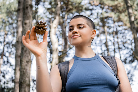 Low angle view of young tattooed and short haired female tourist with backpack holding pine cone...