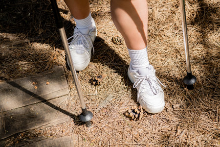 Cropped view of young woman hiker in sneakers standing near trekking poles and pine cones on ground...