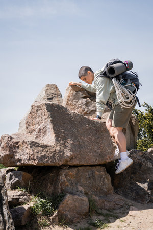 Side view of young short haired female tourist with backpack and travel equipment climbing on stones with blue sky and nature at background, exploring new horizons, summerの写真素材
