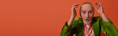 dazed senior man with grey hair, beard and bulging eyes holding hands near head and looking at...